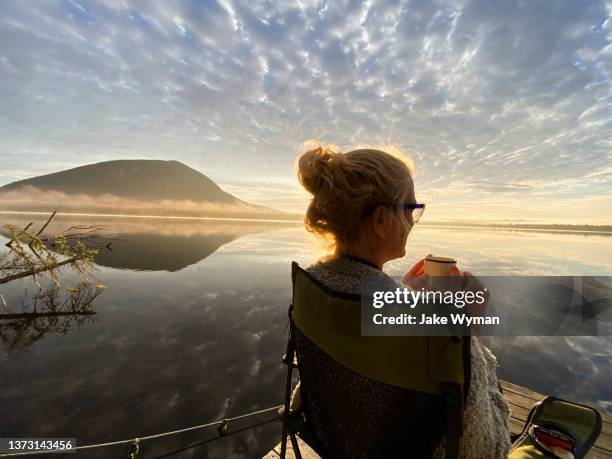 woman sitting in a chair with a hot beverage beside a remote lake in northern maine. - northern maine stock pictures, royalty-free photos & images