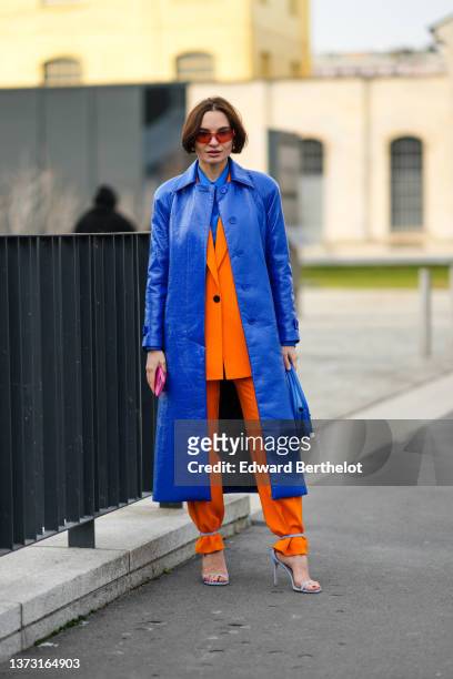Guest wears orange sunglasses, a royal blue electric shirt, a royal blue electric shiny leather long coat, a matching royal blue electric handbag, a...