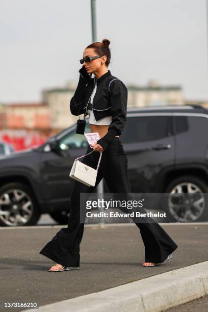 Guest wears black sunglasses, silver earrings, a white with a black strap print pattern cropped top, a black zipper cropped jacket, a white leather...