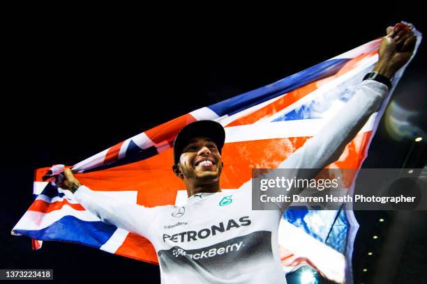 Wearing his logo adorned fire protection suit racing driver overalls undershirt while holding a Union Jack flag above his head, smiling British...