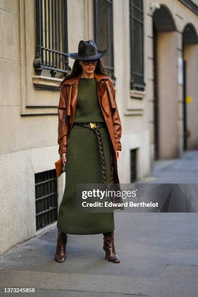 Guest wears a black braided wicker cow-boy hat, brown sunglasses, brown earrings, a dark green ribbed wool long dress, a camel shiny leather long...