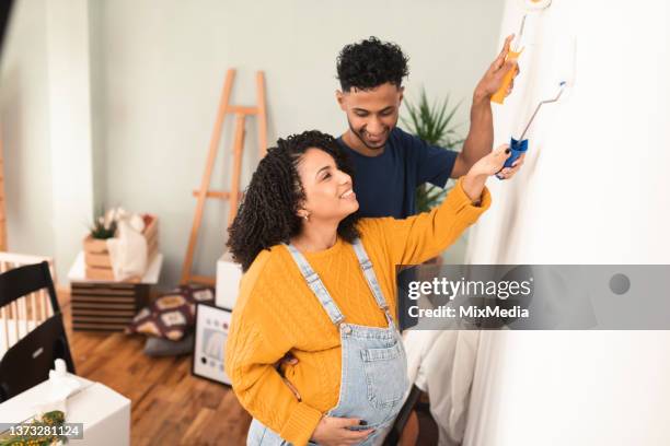 happy young couple expecting a baby and painting the walls at their new home - baby paint stock pictures, royalty-free photos & images