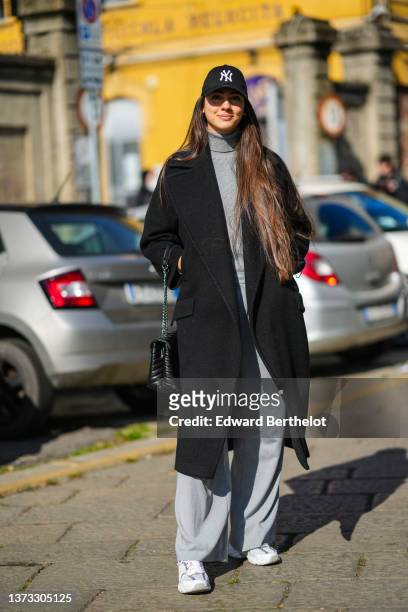 Guest wears a black with white embroidered logo cap from New Era, a pale gray turtleneck pullover, a black oversized long coat, a black shiny leather...