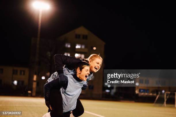 cheerful female athlete piggybacking player on soccer field at night - womens soccer stockfoto's en -beelden