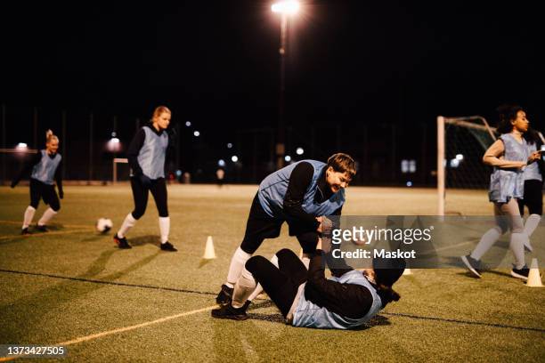 smiling sportswoman helping friend while female athletes practicing on field at night - local soccer field stock pictures, royalty-free photos & images