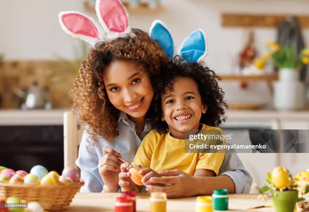 Happy african american family: mother teaching happy little kid soon to decorate Easter eggs while sitting in kitchen