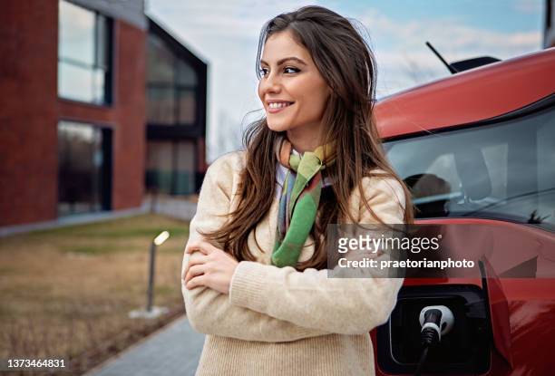 porträt einer jungen frau, die vor ihrem haus ein elektroauto auflädt - autobatterie stock-fotos und bilder