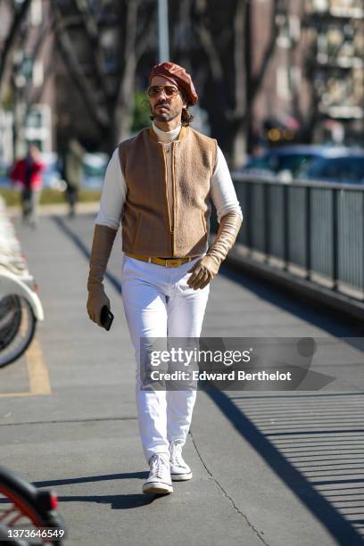 Guest wears a camel shiny leather beret, a white turtleneck pullover, a beige fluffy sleeveless zipper jacket, beige shiny leather high gloves, a...