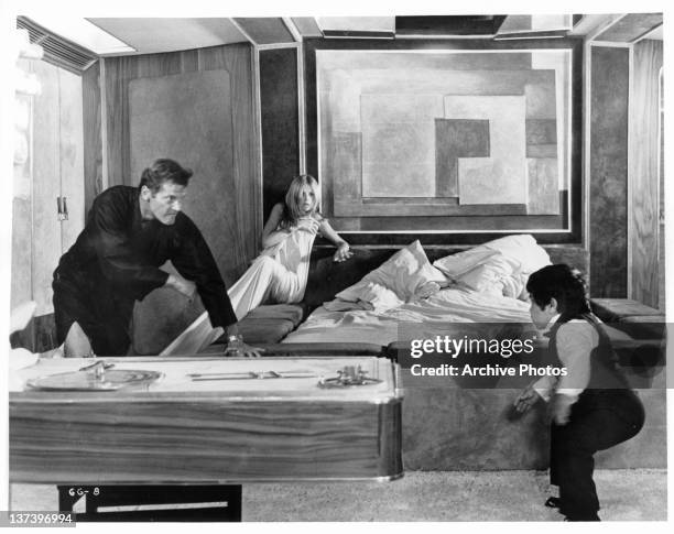 Roger Moore chasing Hervé Villechaize while Britt Ekland huddles against the wall with a sheet wrapped around her in a scene from the film 'The Man...