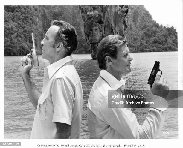 Christopher Lee back to back with Roger Moore, each holding a gun in the air in a scene from the film 'The Man With The Golden Gun', 1974.