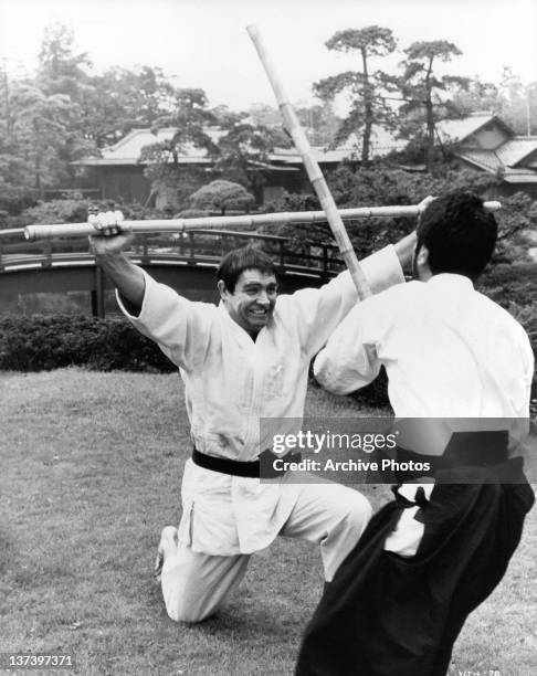 Sean Connery on one knee holding a bamboo stick over his head and wearing martial arts clothing, unknown actor is moving forward to strike him with a...