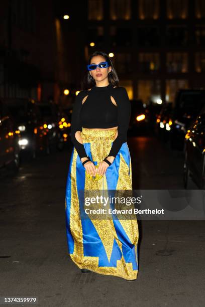 Samia Laaboudi wears blue sunglasses, a black cut-out pattern high neck long sleeves t-shirt, a golf and black small print pattern with royal blue...