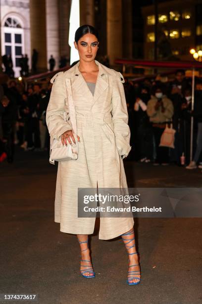 Fiona Zanetti wears a gray shoulder-off dress, a beige oversized belted long coat, a white embossed striped pattern shoulder bag, gold rings, blue...