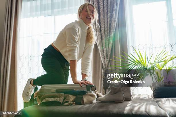 middle age blonde smiling woman in white sweater trying to close suitcase full of clothes in living room at home. female sit on a suitcase and laughing - full stock-fotos und bilder