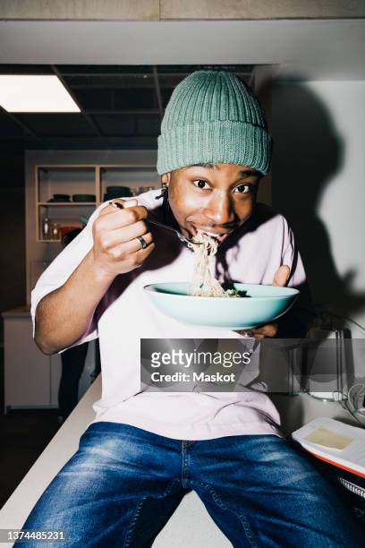 portrait of young man eating noodles while sitting on dining table at college dorm - dorm room stock-fotos und bilder