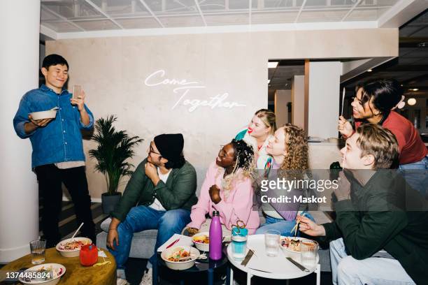 smiling young man photographing multiracial friends holding props while having food in dorm - college dorm party stock-fotos und bilder