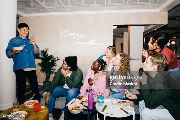 smiling young man photographing multiracial friends holding props while having food in dorm - campus party fotografías e imágenes de stock