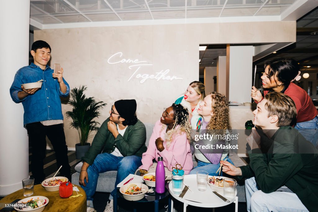 Smiling young man photographing multiracial friends holding props while having food in dorm