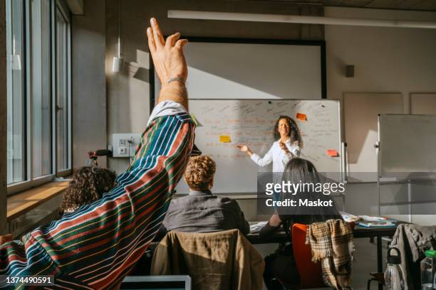 cropped raised hand of male student with friends and teacher in classroom - lernender stock-fotos und bilder