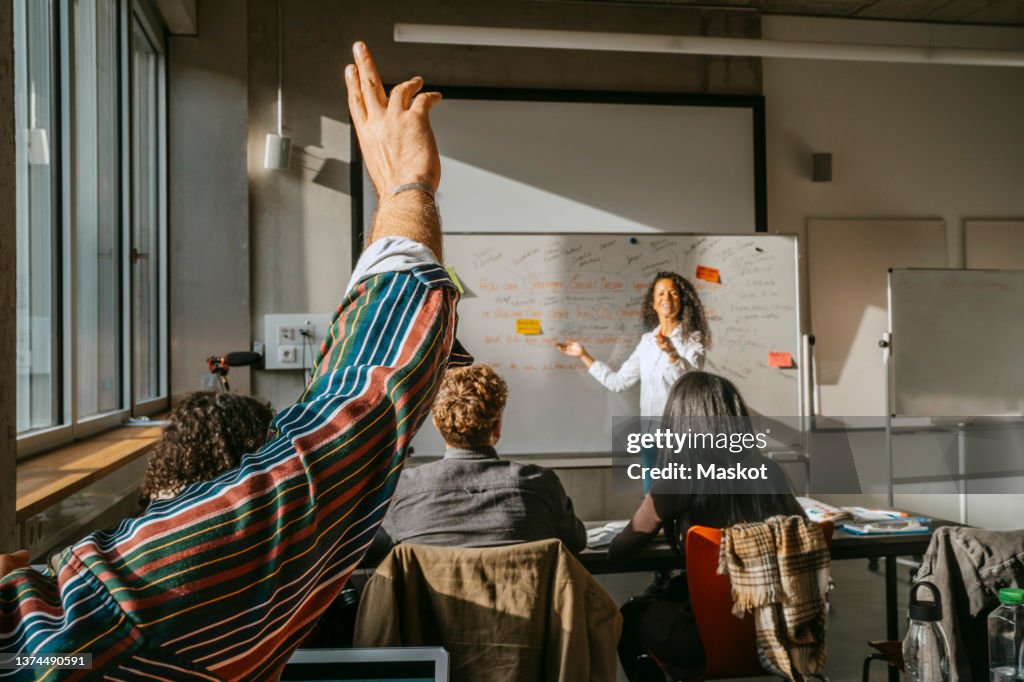 Cropped raised hand of male student with friends and teacher in classroom