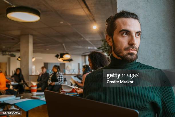 thoughtful young bearded man sitting with laptop in university cafeteria - mature student stockfoto's en -beelden