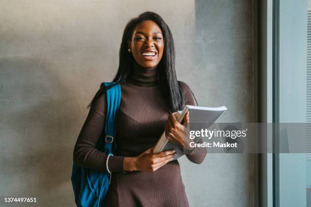 portrait of cheerful young woman holding book standing with backpack against gray wall - portrait student stock pictures, royalty-free photos & images