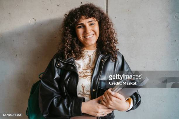 portrait of smiling young woman holding book and backpack leaning on gray wall - studentessa di scuola secondaria foto e immagini stock