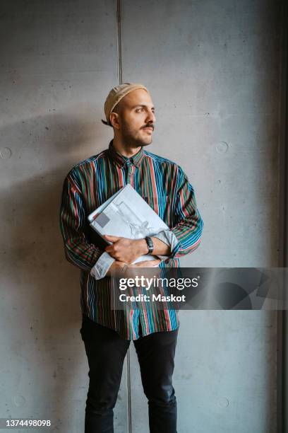 thoughtful male adult student holding book looking away in front of gray wall at college - three quarter length stock pictures, royalty-free photos & images
