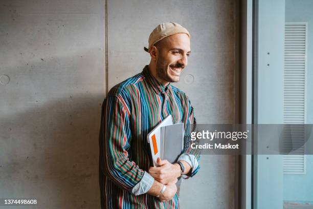 cheerful male adult student holding book standing in front of gray wall at college - student stock-fotos und bilder