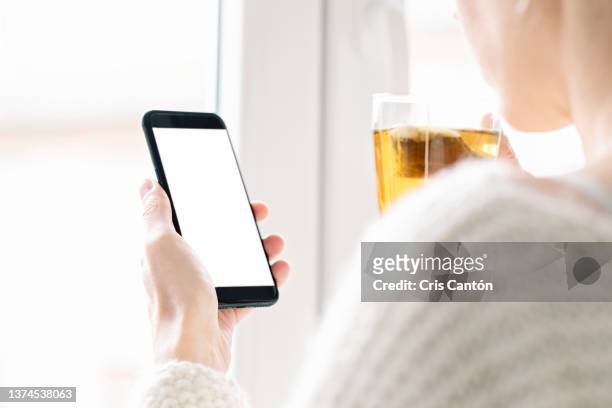 woman holding smartphone with white screen at home - rear view hand window stock pictures, royalty-free photos & images