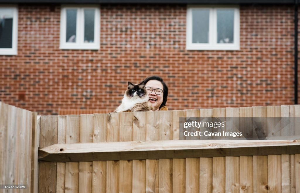 Woman with her cat smiling over garden fence saying hello to neighbour