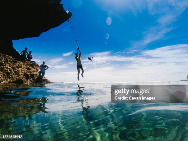 teenage boy swings on rope across teal blue water in summer - altalena di corda foto e immagini stock