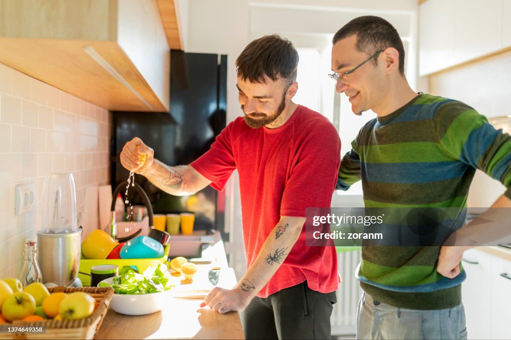 Bearded young man squeezing lemon juice over lettuce in bowl, his partner embracing him