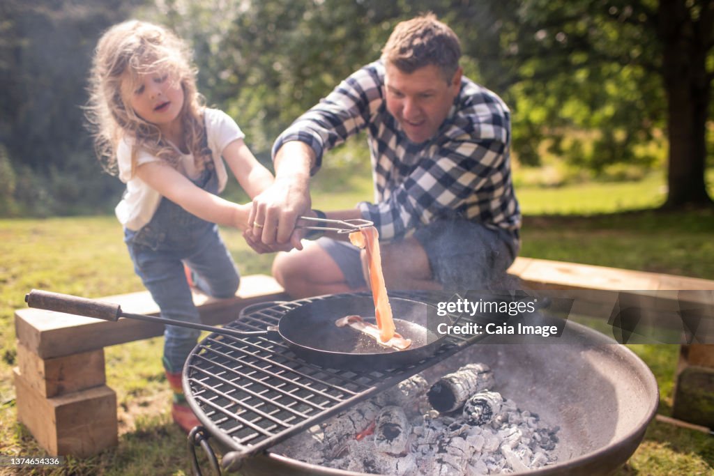 Father and daughter cooking bacon over charcoal grill outdoors