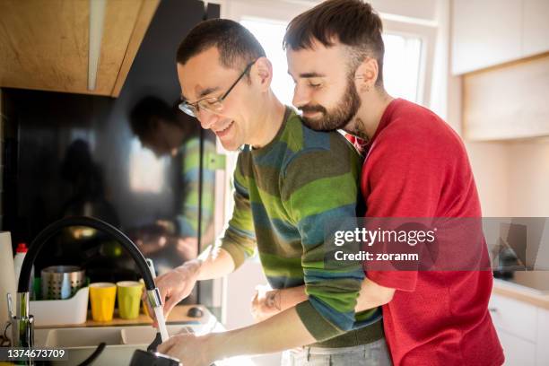 bearded young man hugging his boyfriend from behind, washing up after meal - gay couple in love stock pictures, royalty-free photos & images