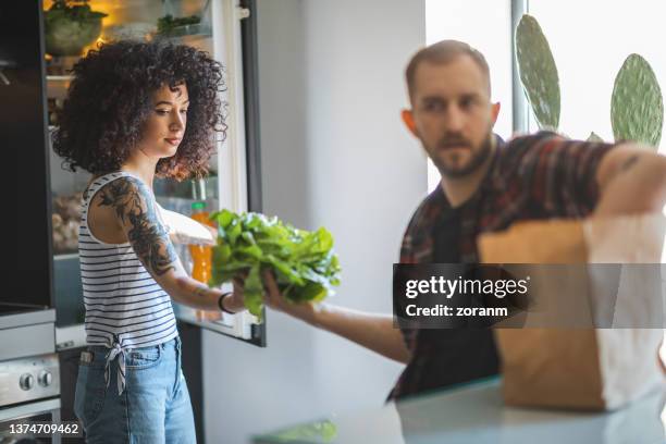 young tattooed woman with curly hair taking lettuce from her friend to put in the fridge - young man groceries kitchen stock pictures, royalty-free photos & images