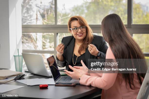 two latina women chat happily as they work on their laptops, sitting at a desk on an ordinary office day. - advice foto e immagini stock