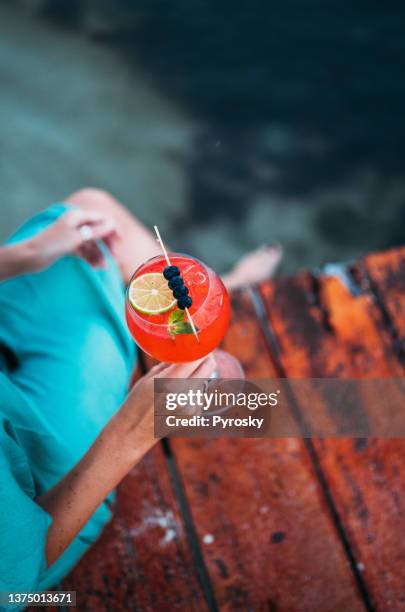 a woman relaxes on a wooden pier with a glass of red cocktail - wood pier stock pictures, royalty-free photos & images