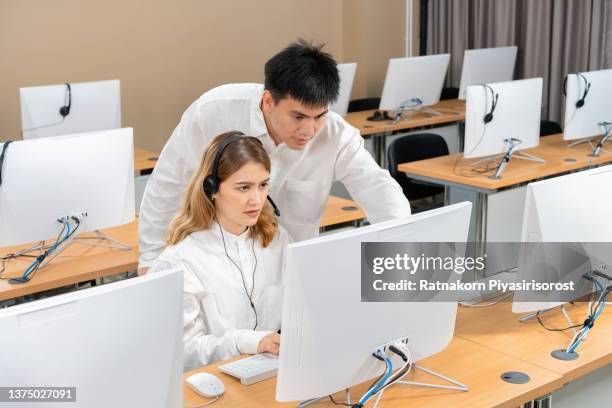 call center employees wearing headset using computer, male manager explains show to new colleague marketing database program, busy service phone operators sitting at shared desk, assistance teamwork concept - internship marketing stock-fotos und bilder