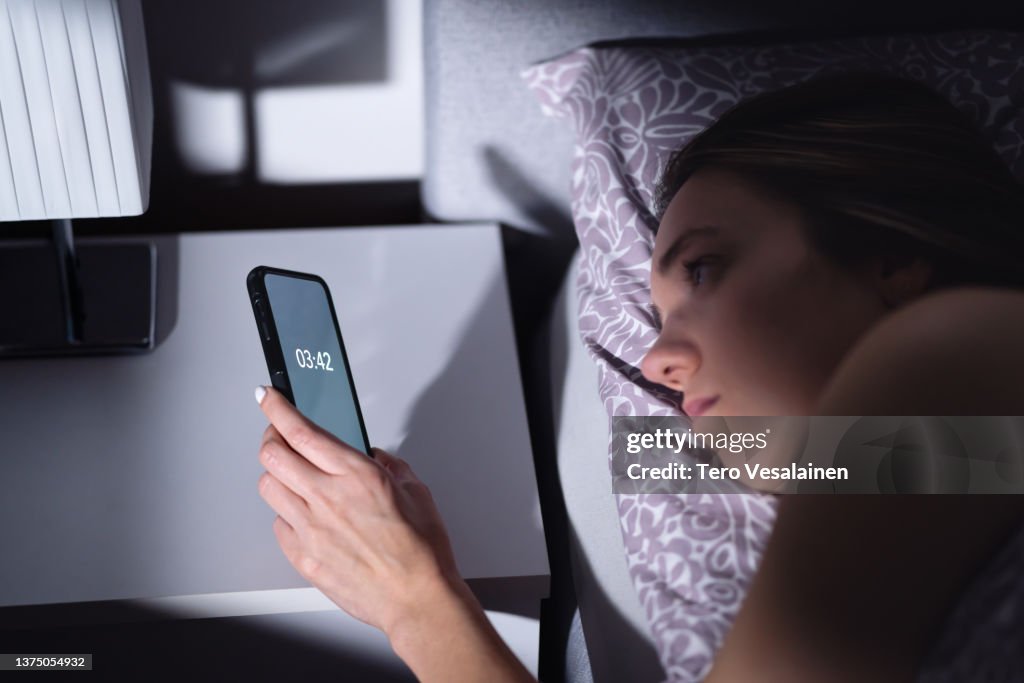 Schlaflosigkeit. Schlaflose Frau, die die Zeit sucht und nachts nicht schlafen kann. Traurige müde Person mit Störung im Bett. Telefonuhr. Schlechter Traum und Problem, wach zu bleiben.