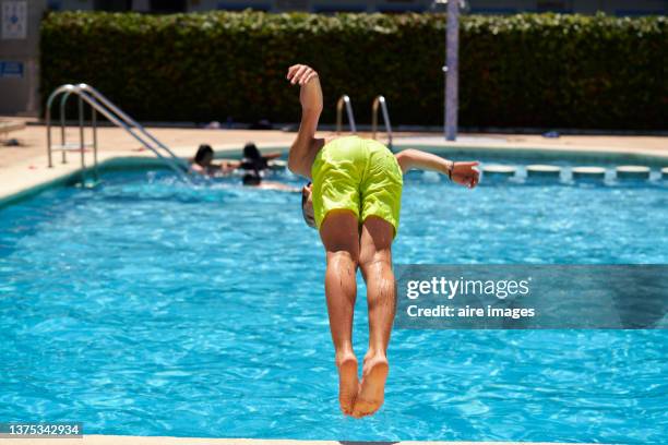 front view of an excited boy in yellow shorts jumps into the hotel pool doing a somersault enjoying of his vacation summer against a group of people blurred background - calções azuis - fotografias e filmes do acervo