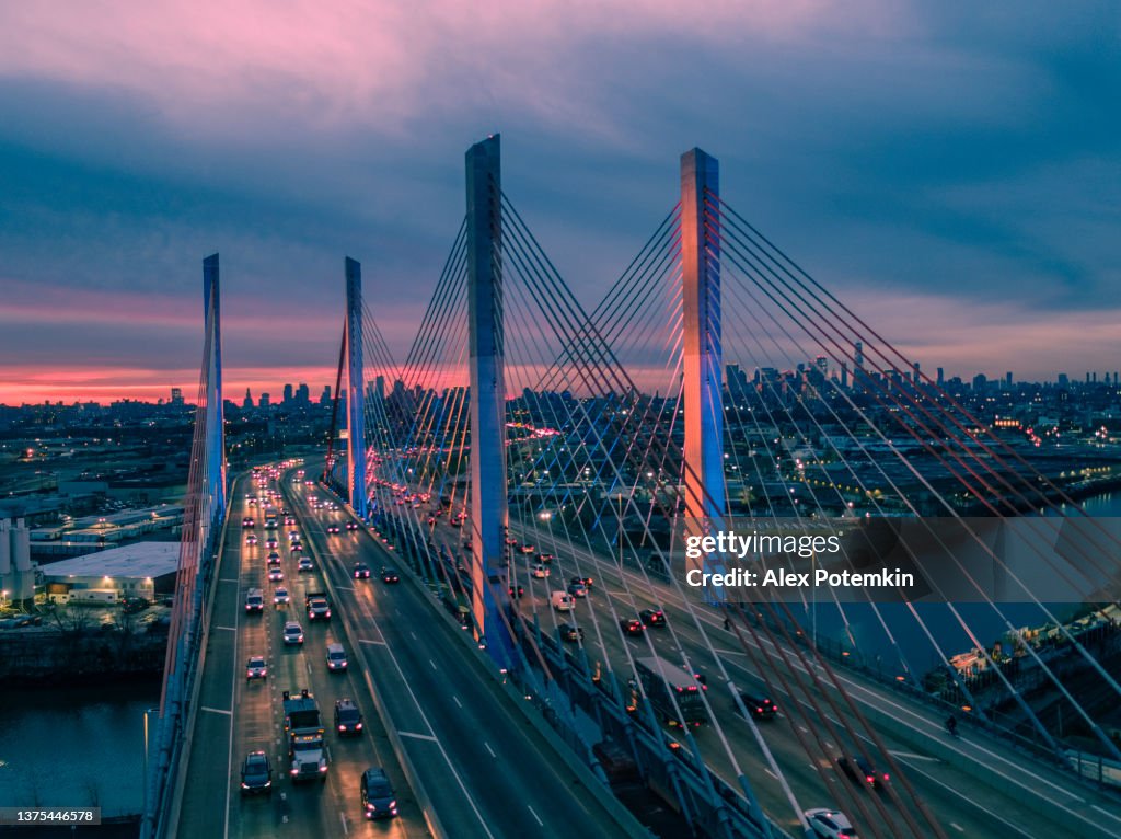 Aerial view of illuminated Kosciuszko Bridge at dusk, with the remote view of Manhattan and Brooklyn over the industrial zone of Williamsburg.