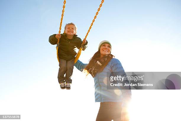mom pushing son on a swing set. - zwaaien activiteit stockfoto's en -beelden