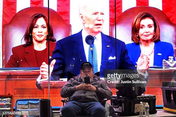 News crew member sits in front of a live feed of U.S. President Joe Biden delivering his State of the Union address that is playing on a big screen...