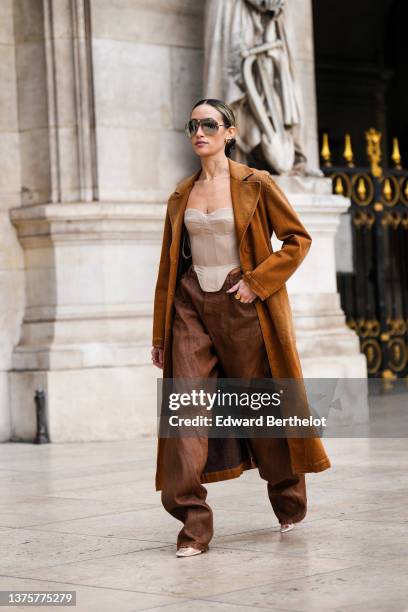 Alexandra Guerain wears gold earrings from Louis Vuitton, black sunglasses, a gold chain pendants necklace, a brown camel denim long coat, a beige...