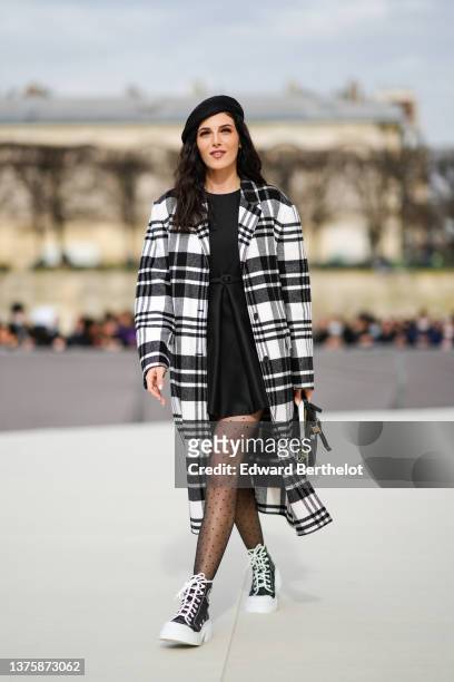 Razane Jammal wears a black felt / wool beret hat, a black pleated short dress with a black satin small belt from Dior, a black and white checkered...