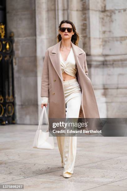 Chloe Lecareux wears brown sunglasses, a white gold silk wrap / knot cropped blouse, a beige long coat, matching white gold silk large pants, a white...