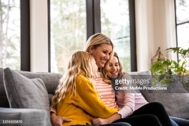 mother and tween daughters embracing on couch in modern home - family hugging bright stock pictures, royalty-free photos & images