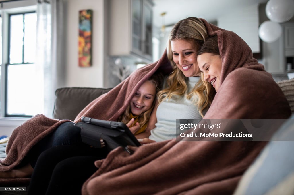 Mother and tween daughters snuggled under blanket and watching a movie on digital tablet
