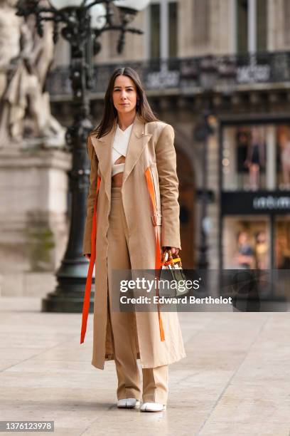 Alice Barbier wears a white cross-neck cropped top, a beige long coat with neon orange straps, high waist beige wide legs flared pants, a white shiny...